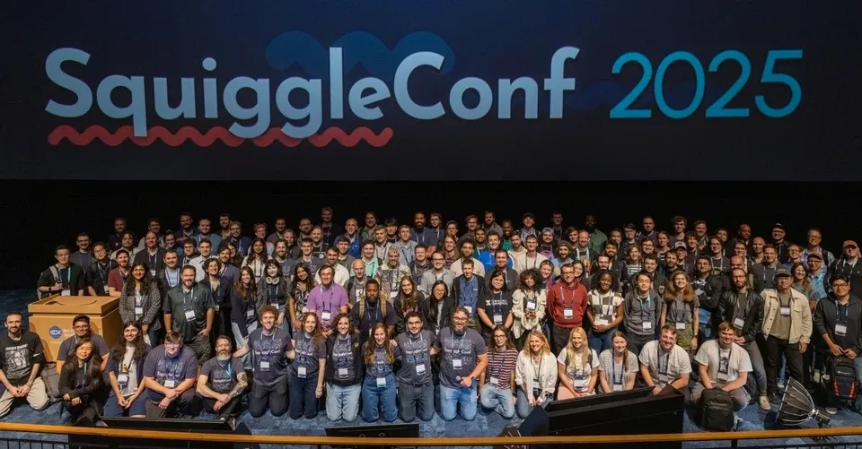 High-level photo of everyone kneeling or standing, smiling, in the front of an auditorium. On the screen, the SquiggleConf 2025 logo is visible.